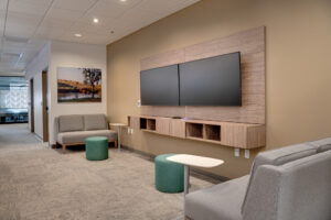 Flexible working space with two TV monitors on side wall with two small loveseat couches and work tables besides.