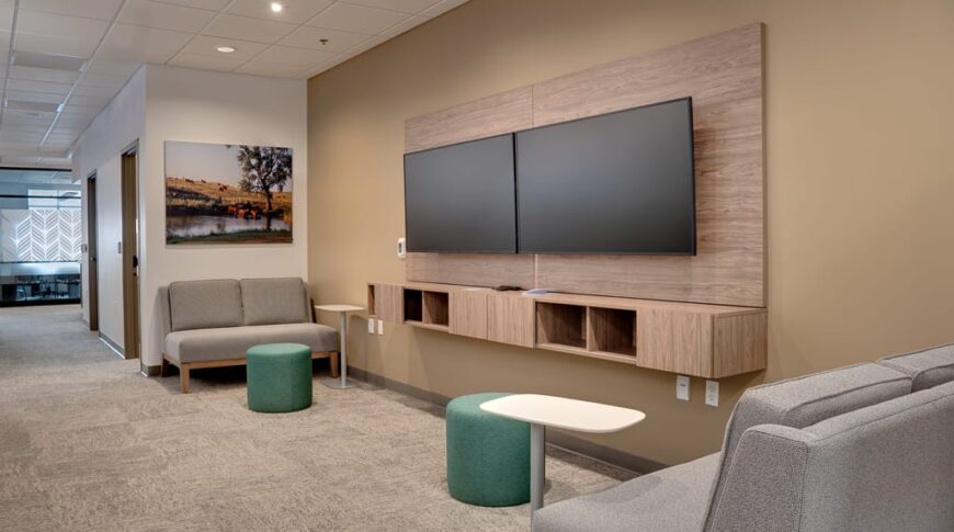 Flexible working space with two TV monitors on side wall with two small loveseat couches and work tables besides.