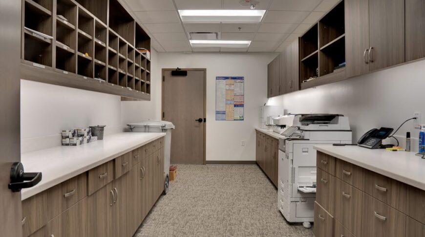 Printing workroom with upper and lower cabinetry on either side and copying machine and other office equipment and supplies throughout.