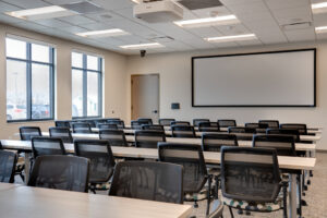 Large meeting room with classroom style tables and chairs looking forward to large projector screen. Windows on left hand side of room.