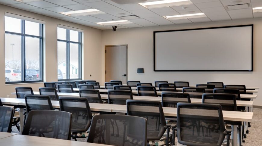 Large meeting room with classroom style tables and chairs looking forward to large projector screen. Windows on left hand side of room.