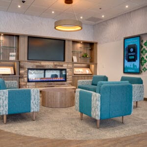 Front waiting area of AgCountry Farm Credit Services in Lamoure, ND, with brick facade electric fireplace and cabinetry flanking each side, aqua blue seating chairs and coffee tables in front, and electronic interactive display monitor to the side.