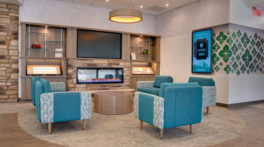 Front waiting area of AgCountry Farm Credit Services in Lamoure, ND, with brick facade electric fireplace and cabinetry flanking each side, aqua blue seating chairs and coffee tables in front, and electronic interactive display monitor to the side.
