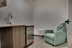 Mother's room equipped with nursing rocking chair, side table, and convenience wash station with minifridge besides.