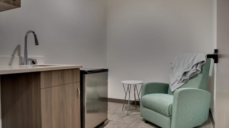 Mother's room equipped with nursing rocking chair, side table, and convenience wash station with minifridge besides.