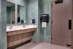 Bathroom interior view of wood-grained laminate vanities and door, with light green tiles on every wall from floor to ceiling.