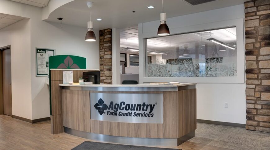 AgCountry Farm Credit Services front reception desk with curved wood laminate and company sign on metal logo over the laminate. Stone columns flank each side of the desk.