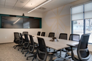Modern conference room with light walls and carpeting, along with long, light wood-colored conference table surrounded by black chairs overlooking double monitors on back wall.
