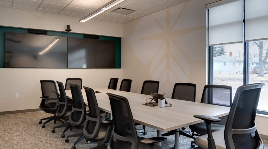 Modern conference room with light walls and carpeting, along with long, light wood-colored conference table surrounded by black chairs overlooking double monitors on back wall.