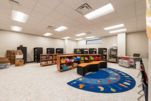 Small school library with rows of library shelves on the back two walls and some shorter bookshelves in the middle of the room besides a learning mat.