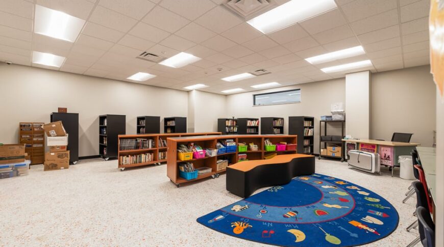 Small school library with rows of library shelves on the back two walls and some shorter bookshelves in the middle of the room besides a learning mat.