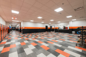 School commons area with large checkered floor in the school colors, white, orange, and gray. Kitchen pass-through window in the back with hallway of lockers down the left side.