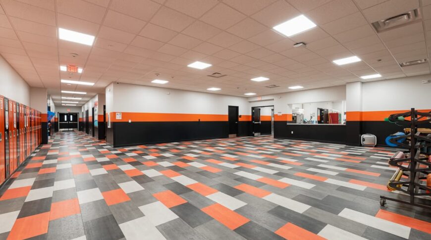 School commons area with large checkered floor in the school colors, white, orange, and gray. Kitchen pass-through window in the back with hallway of lockers down the left side.