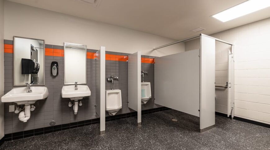 Newly constructed school boys bathroom with gray flooring and gray and white walls with an orange accent band.