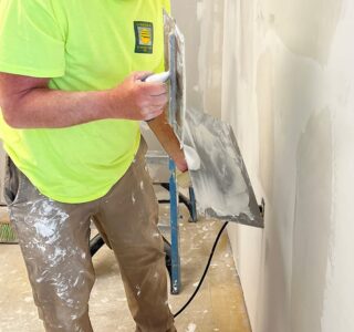 Old man with paint-splattered khaki pants and yellow safety shirt wearing baseball hat while he's mudding drywall.