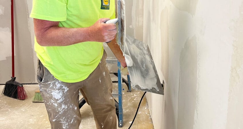 Old man with paint-splattered khaki pants and yellow safety shirt wearing baseball hat while he's mudding drywall.