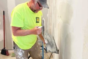 Old man with paint-splattered khaki pants and yellow safety shirt wearing baseball hat while he's mudding drywall.