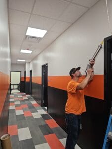 Man with orange safety shirt and hat reaching up to calk around a newly installed door frame at a school.