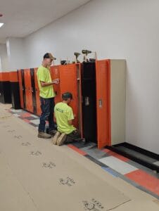 Two young men with jeans and white safety shirts and baseball hats installing orange and black lockers at a school.