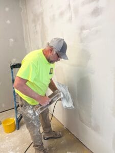 Old man with paint-splattered khaki pants and yellow safety shirt wearing baseball hat while he's mudding drywall.
