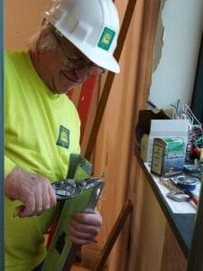 Old man with white hair and glasses wearing white hard hat and yellow long-sleeve safety shirt using tool to bend metal. Other repair equipment in background.