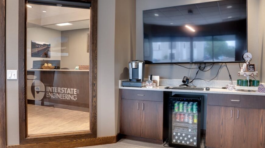 Convenience station with cabinetry, minifridge, and coffee maker. TV overhead. Floor to ceiling window to the left overlooking the reception desk for Interstate Engineering.