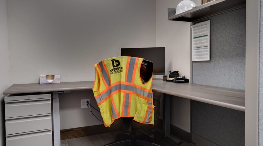 Office cubicle in standard gray with lower file cabinets and one upper shelving. Computer monitor and chair besides with a safety vest that says "Interstate Engineering" on it.