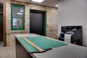Workroom with long terrazzo island with green cutting mat and ruler on top. Printer in back right. Original brick wall and green-framed window to the back.