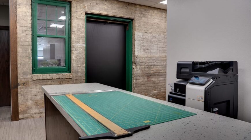 Workroom with long terrazzo island with green cutting mat and ruler on top. Printer in back right. Original brick wall and green-framed window to the back.
