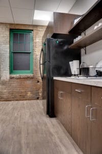 Kitchenette cabinetry and open upper shelving with Black fridge. Behind all is an original brick building wall and window trimmed in green.