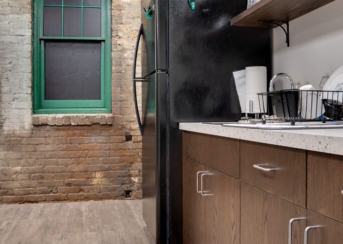 Kitchenette cabinetry and open upper shelving with Black fridge. Behind all is an original brick building wall and window trimmed in green.