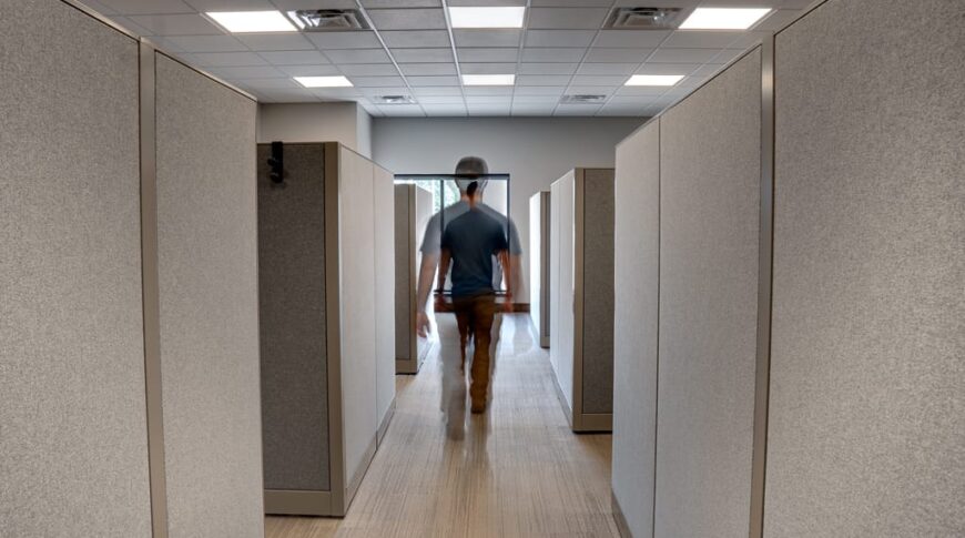 Blue-collar man walking down aisle of tall cubicles.