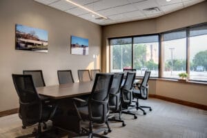 Conference room with long table and black chairs surrounding. Wall of windows to back right, and project pictures displayed on left wall.