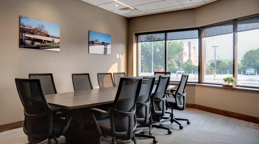 Conference room with long table and black chairs surrounding. Wall of windows to back right, and project pictures displayed on left wall.