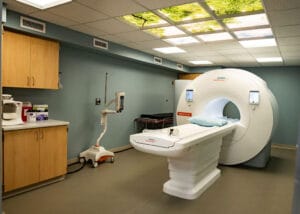 MRI room with MRI machine in the middle and overhead fluorescent lights with treetop visual inserts. Cupboard station to the left.