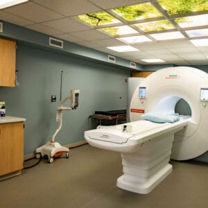 MRI room with MRI machine in the middle and overhead fluorescent lights with treetop visual inserts. Cupboard station to the left.
