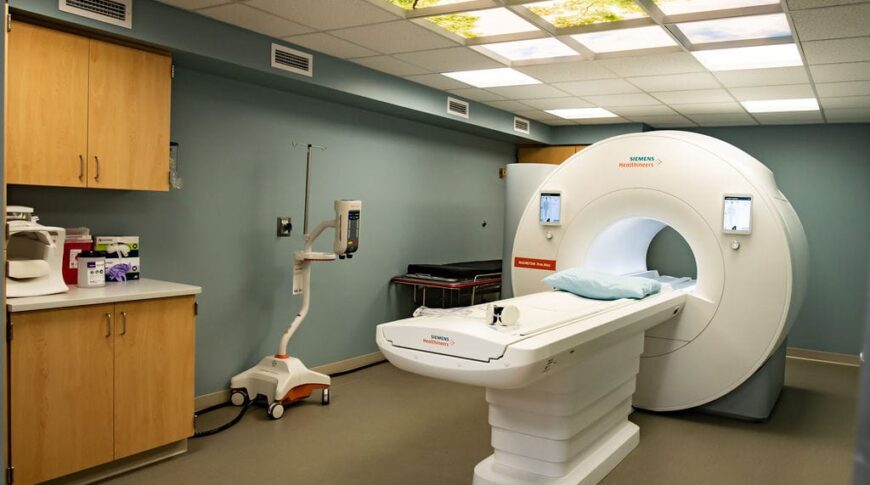 MRI room with MRI machine in the middle and overhead fluorescent lights with treetop visual inserts. Cupboard station to the left.
