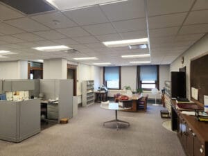 Office interior space with cubicle to the left, open space in center, and office surrounding. Dropped ceiling and carpet surrounded by beige walls.