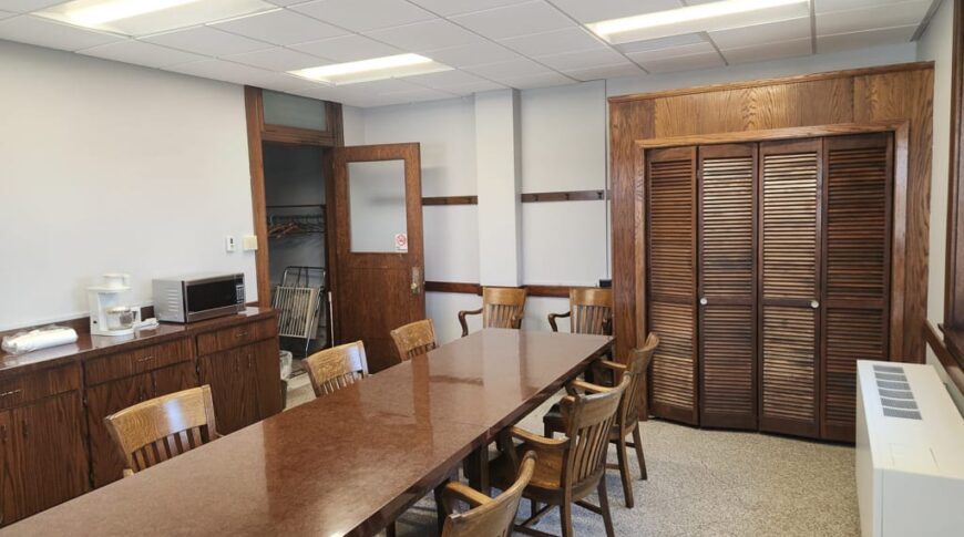 Conference room with long rectangular wooden table with wooden chairs surrounding and wooden bifold closet doors in the background.
