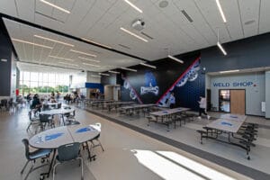 Interior common area of CTE center with Dragons school mascot decal on the back wall. Cafeteria tables and students throughout the space. Photo by Icon Architectural Group.