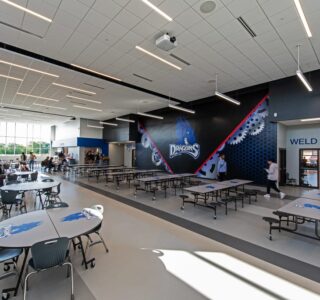 Interior common area of CTE center with Dragons school mascot decal on the back wall. Cafeteria tables and students throughout the space. Photo by Icon Architectural Group.