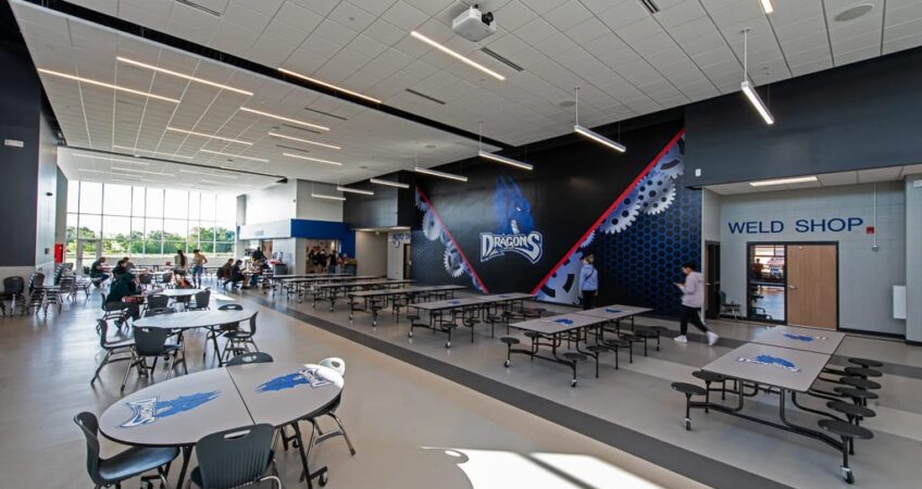 Interior common area of CTE center with Dragons school mascot decal on the back wall. Cafeteria tables and students throughout the space. Photo by Icon Architectural Group.