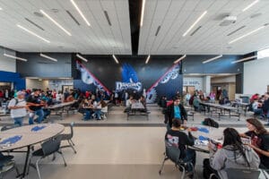 School commons area with foldable cafeteria tables throughout, accent wall with dragon mascot in the background, and accent lighting above. Photo by Icon Architectural Group.