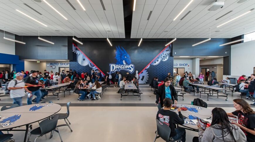 School commons area with foldable cafeteria tables throughout, accent wall with dragon mascot in the background, and accent lighting above. Photo by Icon Architectural Group.
