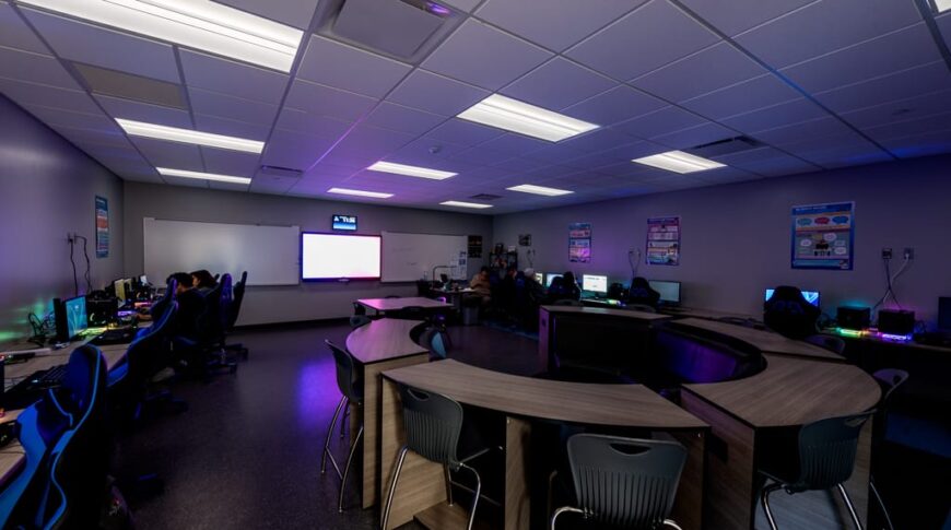 Dimly lit computer lab with desks around the perimeter of the room and a gathering pod in the center. Photo by Icon Architectural Group.