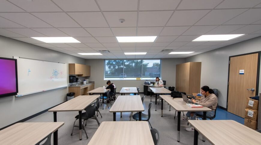 Classroom view from the side looking down long light colored desks with whiteboard at the far left and windows to the back wall. Photo by Icon Architectural Group.