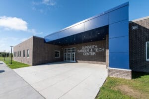 Exterior of dark brown brick school with blue architectural panels. A metal sign that says "Dunseith Career & Tech Center" by the entrance doors. Photo by Icon Architectural Group.