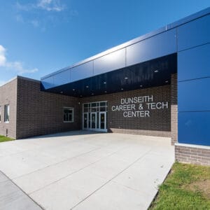 Exterior of dark brown brick school with blue architectural panels. A metal sign that says "Dunseith Career & Tech Center" by the entrance doors. Photo by Icon Architectural Group.