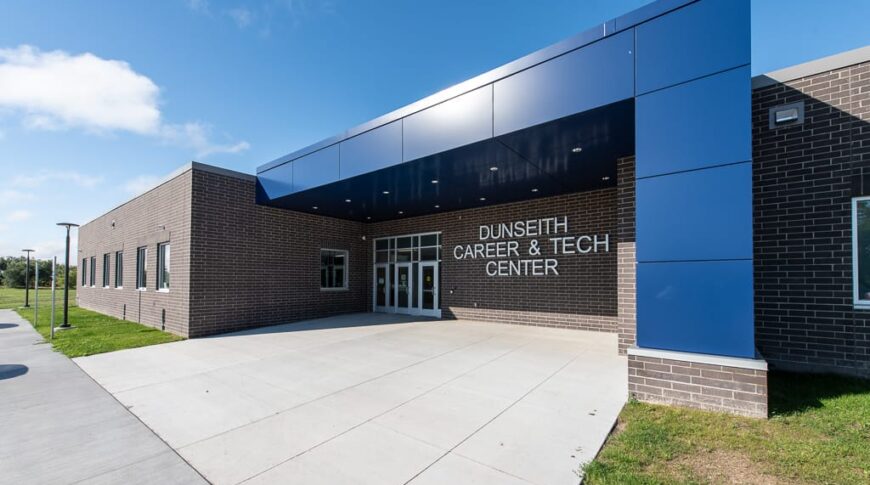 Exterior of dark brown brick school with blue architectural panels. A metal sign that says "Dunseith Career & Tech Center" by the entrance doors. Photo by Icon Architectural Group.