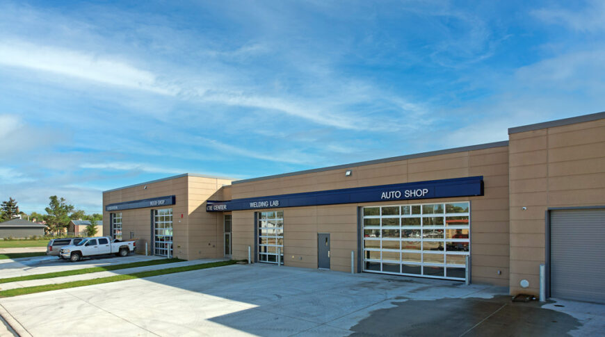 Exterior of CTE school with four large garage bays with full-window doors and a blue ribbon above each with a label for which shop it is. Photo credit to Icon Architectural Group.